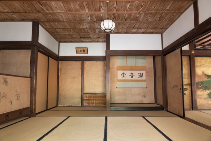 A tatami-floored spacious room is seen from a low angle, with the right half of the back wall as an alcove displayiing a hanging scroll of calligraphy.