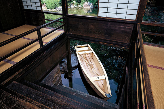 An anchored, wooden boat of the size for a couple of people is seen below through the water entrance from the inside of the raised-floor of the pavilion.