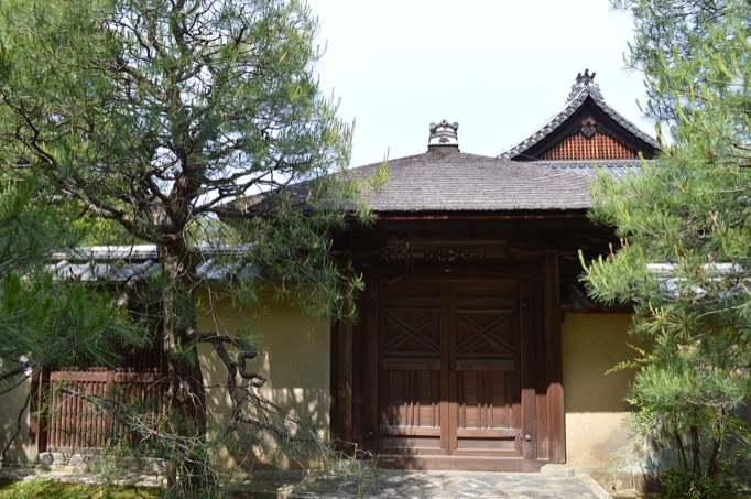 A roofed gate with its doors closed is seen in between pine trees. Seen behind the gate is the top of the gable roof of the temple building.