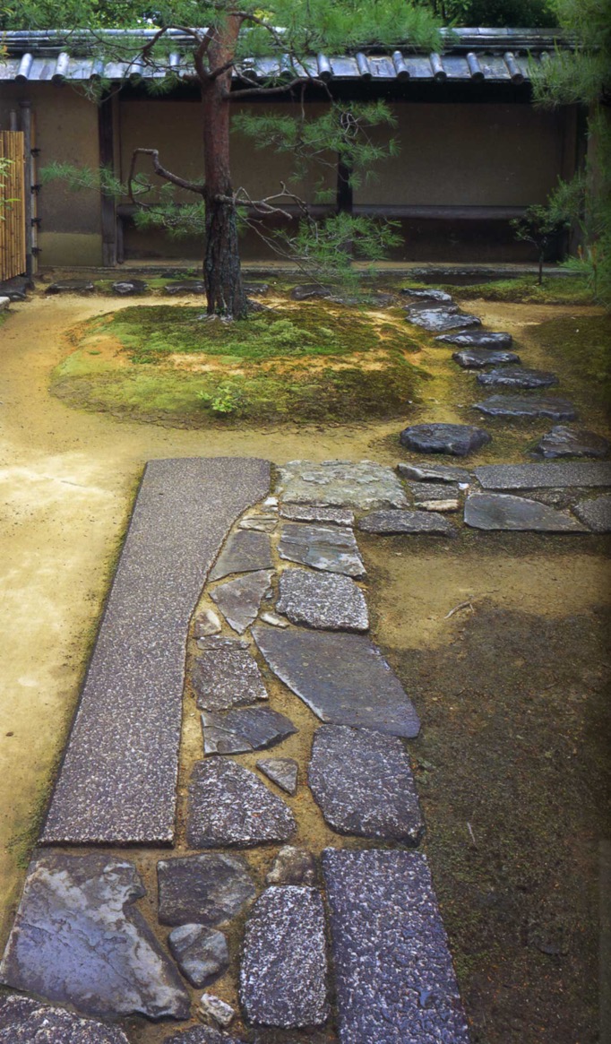 Flat-top stones are laid out to form a right-angled footpath, turning to the right in front of the pine tree behind which a roofed bench is seen.
