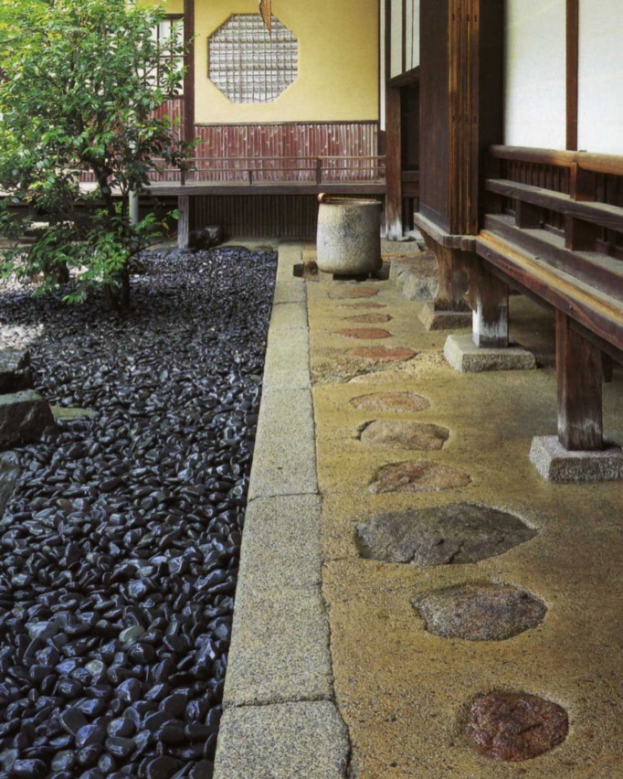 A straight sequence of flat-top stones, embedded in the rectangular tataki floor, leads to a cylindrical stone furniture with a wooden ladle on its top. The dark blue gray pebbles are packed along the straight edge of the tataki floor on the left.