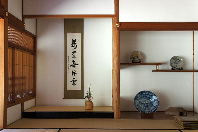 A tatami-floored room with its backwall partitioned into the wood-floored alcove on the right and the tatami-floored alcove on the left, the latter of which has a tie beam at a higher position and displays a hanging scroll of calligraphy.