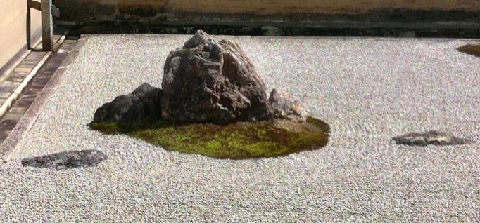 A large rough-looking rock in the middle, flanked by a pair of smaller rocks, all of which sit on a patch of moss. One flat-top rock embedded in the gravel ground to the left of the moss patch, and another to the right.